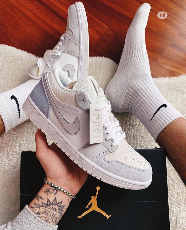 White sneakers with a logo held by a person wearing white socks and gray pants.