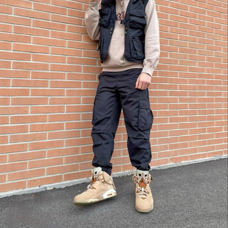 Person wearing a beige hoodie, black vest, black pants, and tan boots against a brick wall.