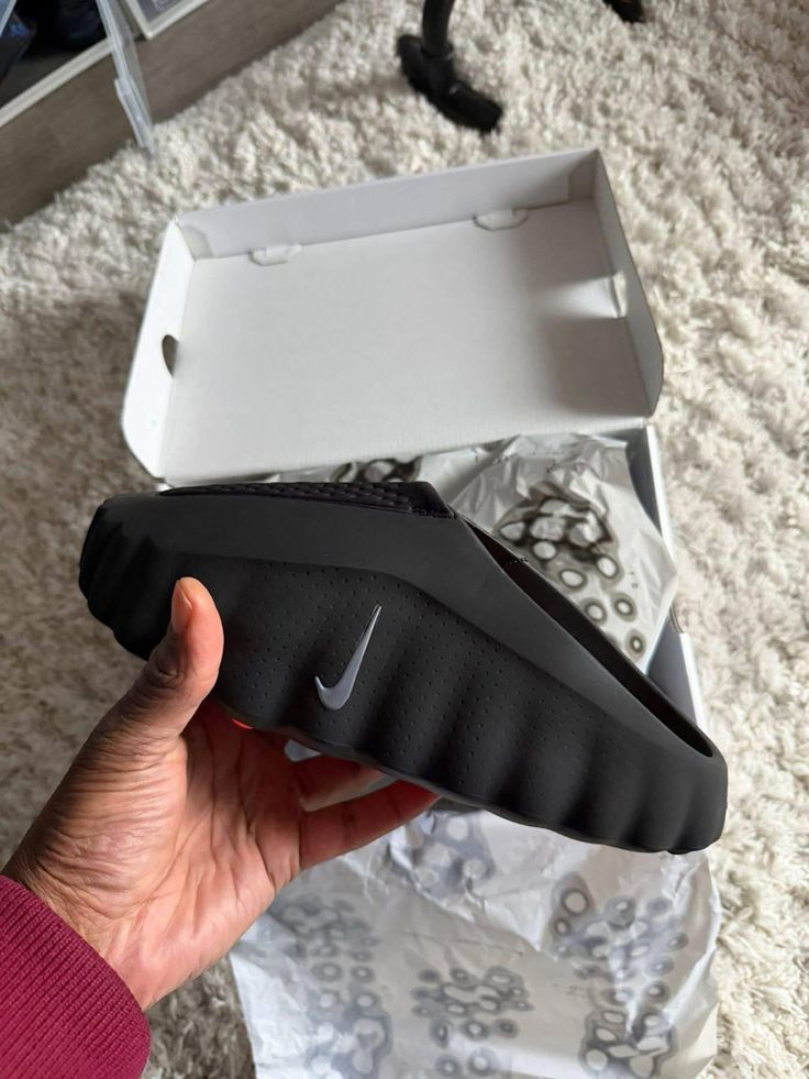 Black sandal with a Nike logo held by a hand, with an open white box in the background.